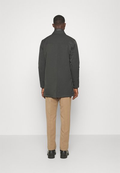 Abrigo largo gris oscuro con corte recto, con un alto cuello y dos bolsillos laterales, combinado con pantalones beige y zapatos negros.