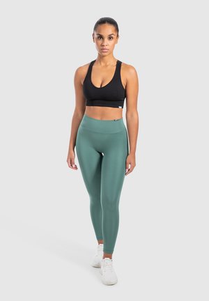 Zwarte geribbelde sport-bh gecombineerd met hooggetailleerde, gladde, saliegroene leggings. Witte sneakers maken de sportoutfit compleet.