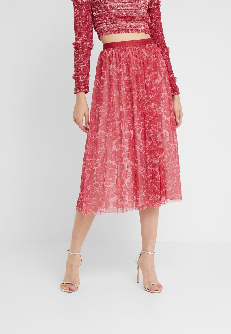 Femme portant une jupe midi rouge fleurie avec un crop top rouge smocké et des sandales argentées à talons hauts avec bride à la cheville, debout sur un sol blanc.