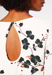 Krämfärgad topp med öppen rygg och detalj i form av ett nyckelhål. Den har ett blommönster i mörkgrön och bruna accenter på tyget.