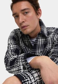 Czarnobiała koszula w kratkę z teksturowanej tkaniny, dużym kołnierzem, zapinanymi mankietami i luźnym krojem. Biała koszulka na krótki rękaw widoczna pod spodem.