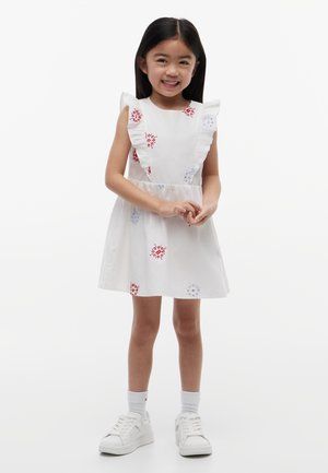 Joven niña sonriendo, vestida con un vestido blanco con estampados florales rojos y azules, mangas con volantes, calcetines blancos y zapatillas blancas, de pie sobre un fondo blanco.