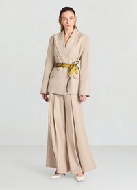 Blazer beige ajusté avec des boutons croisés, associé à un pantalon à jambes larges. Une ceinture foulard jaune à motifs ajoute du détail, avec des talons aiguilles.
