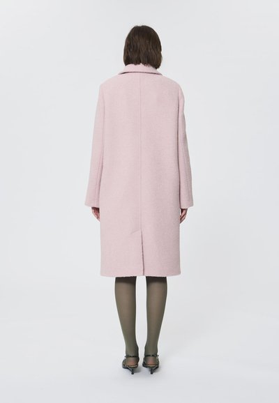 Cappotto in lana rosa con colletto, taglio dritto e maniche lunghe. Presenta una cucitura centrale sul retro e un orlo fino al ginocchio. Indossato con collant olive.