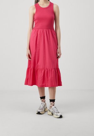 Kvinde iført en ærmeløs, knaldrød midi-kjole med flæsekant, gennemsigtige sorte strømper med hjerteprint og sølv sneakers, stående på en ensfarvet baggrund.