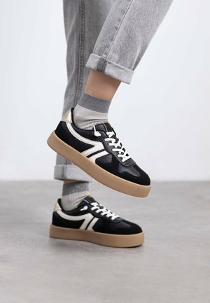 Zapatillas en blanco y negro con un diseño texturizado, que presentan una suela de goma, cordones en la parte delantera y acentos en contraste. Jeans grises enrollados y calcetines visibles.