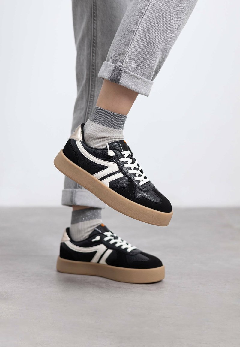 Zapatillas en blanco y negro con un diseño texturizado, que presentan una suela de goma, cordones en la parte delantera y acentos en contraste. Jeans grises enrollados y calcetines visibles.