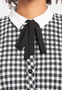 Mustavalkoinen ruudullinen paita, jossa on valkoinen kaulus, musta nauhatiedot kauluksessa ja mustat napit.