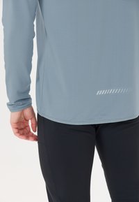 Lyseblå langærmet top med et tekstureret mønster, der har en diskret reflekterende detalje nær kanten, parret med sorte tætsiddende bukser.