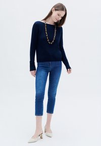 Dunkelblauer Strickpullover mit weitem Ausschnitt, kombiniert mit knöchellangen blauen Jeans und cremefarbenen Blockabsatzschuhen. Langer Kettenanhänger als Akzent.