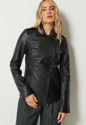 Vrouw met blond haar die een nauwsluitend zwart leren jack draagt met ritszakken en een donkere broek, staand tegen een witte achtergrond.