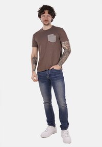 T-shirt marrone a manica corta con collo a giro e tasca testurizzata, abbinato a jeans in denim blu e sneakers bianche. Il modello si presenta in modo informale con tatuaggi visibili.