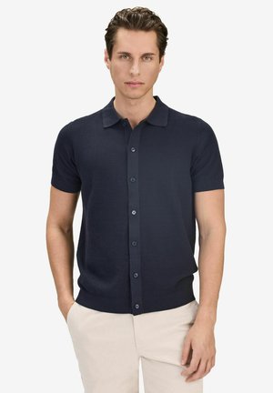 Man draagt een marineblauw poloshirt met korte mouwen en knopen, en lichtbeige broek, staat met handen in de zakken, neutrale gezichtsuitdrukking.
