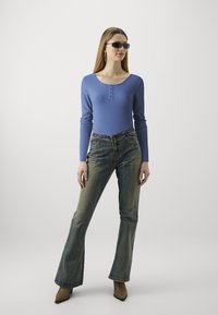 Top blu ribbed a maniche lunghe con dettagli di bottoni, abbinato a jeans a zampa d'elefante in lavaggio scuro e décolleté beige a punta. La modella indossa occhiali da sole neri.