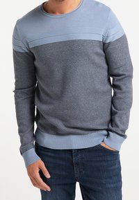 Homme portant un pull en tricot texturé bleu avec des accents bleu clair, associé à un jean bleu foncé, se tenant avec une main dans sa poche.