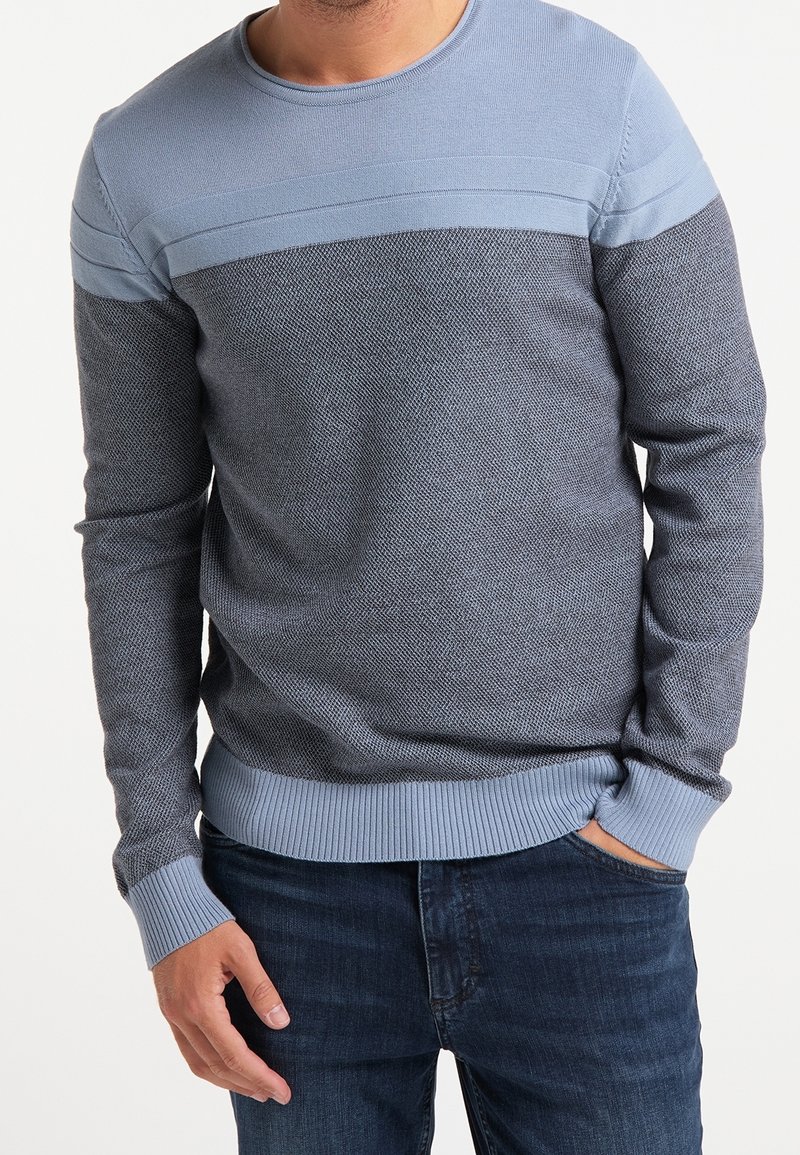 Homme portant un pull en tricot texturé bleu avec des accents bleu clair, associé à un jean bleu foncé, se tenant avec une main dans sa poche.