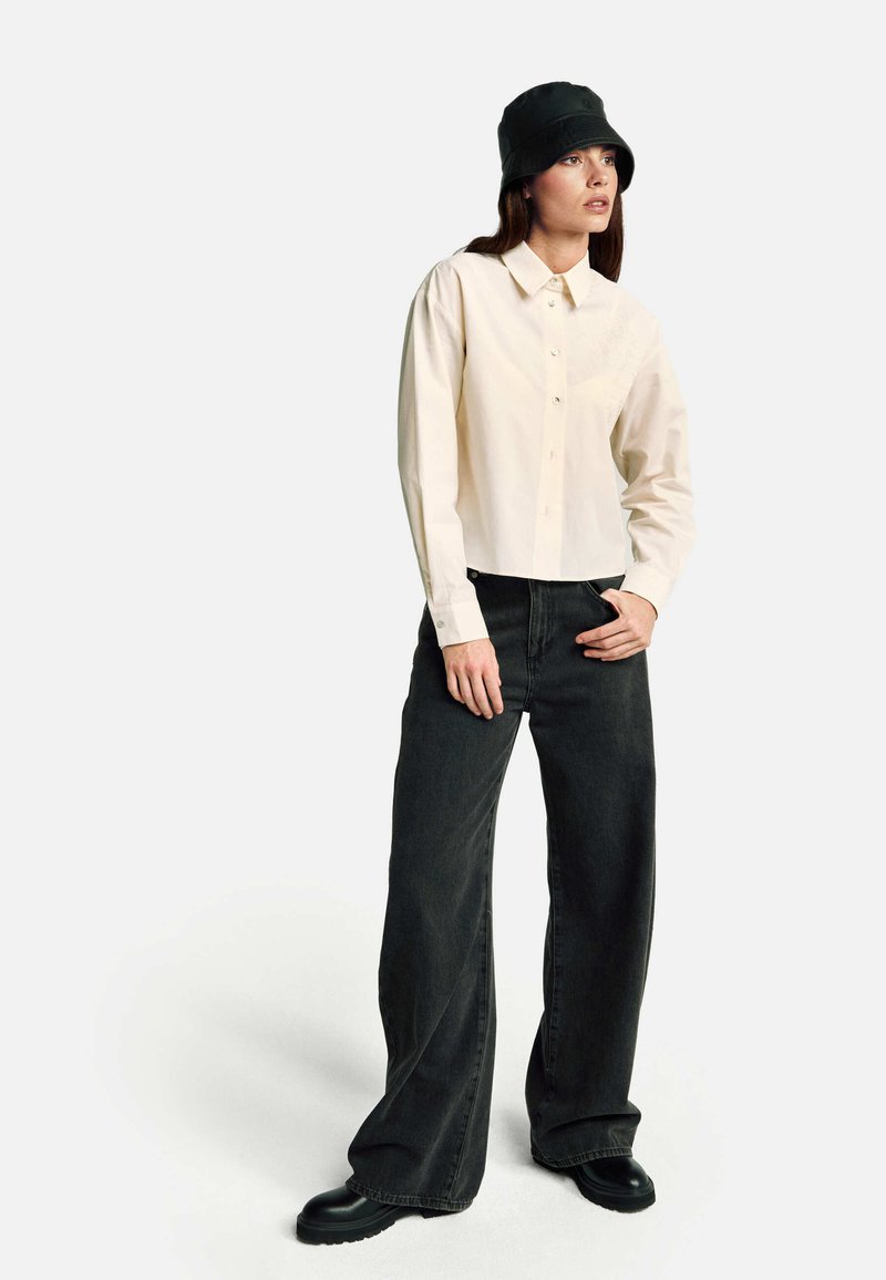 Jasnobeżowa koszula na guziki z długimi rękawami, zestawiona z luźnymi czarnymi jeansami i czarnymi masywnymi butami. Modelka nosi czarny kapelusz typu bucket.