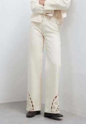 Persona che indossa pantaloni a vita alta color crema a gamba larga con ritagli floreali vicino alle caviglie e scarpe nere, in piedi contro un muro bianco.