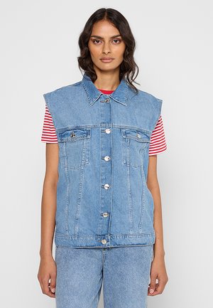 Woman wearing light blue sleeveless denim jacket over red and white striped shirt, paired with light blue denim jeans, standing against white background.