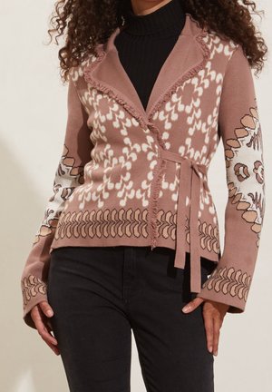Femme portant un pull cache-cœur marron à motifs avec des franges et une ceinture à nouer sur un col roulé noir et un pantalon noir.