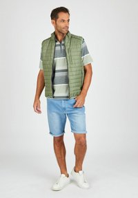Hellgrüne gepolsterte Weste über einem gestreiften kurzärmeligen Hemd, kombiniert mit gekrempelten Jeansshorts und weißen Turnschuhen. Einfaches, lässiges Outfit.