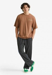 T-shirt di cotone marrone con maniche corte e testo verde, abbinata a pantaloni grigi scuri con righe laterali blu e sneakers verdi.