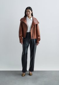 Smeđi kožni skraćeni kaput s širokim ovratnikom, bijela košulja ispod, crne hlače visokog struka i metalik zlatne štikle s oštrim vrhom.