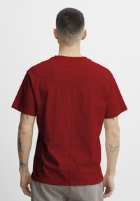 Man with short hair and tattoos on arms facing away, wearing a plain red t-shirt and gray pants against white background.