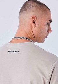 Camiseta de color beige claro con mangas cortas, que presenta un logo negro "PXP" en la espalda. El modelo lleva un collar de cadena plateada. Textura suave.