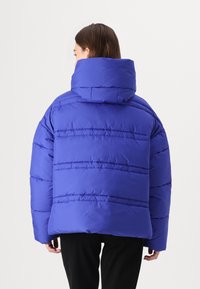 Helle blaue Daunenjacke mit hohem Kragen, die horizontale Nähte aufweist und einen lockeren Sitz hat. Das Material wirkt glatt und gepolstert.