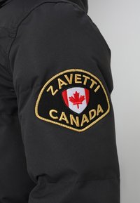 Czarna puchowa kurtka z okrągłym naszywką na rękawie z napisem "ZAVETTI" i "CANADA" w złotym kolorze, obok projektu czerwonego klonu.