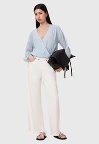 Himmelblaues, strukturiertes Wickeloberteil, weiße weite Jeans mit fransigem Saum und schwarze Tasche; das Modell trägt metallische Sandalen.