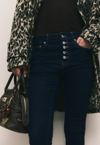 Augsta vidukļa tumši zila džinsu bikses ar piecām pogām priekšpusē, apvienotas ar leopardu raksta mētelīti un teksturētu brūnu topu, turot tumšu somiņu.