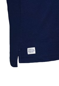 Saum eines dunkelblauen Poloshirts mit weißem rechteckigem Etikett, auf dem "Original Brand U.S. Polo Assn. Since 1890" steht, und einem seitlichen Schlitz.