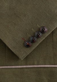Gros plan sur le poignet d'une veste texturée vert olive avec quatre boutons marron foncé et des boutonnières marron sur un tissu ajusté.