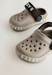 Jasnoszare klapki z otworami wentylacyjnymi, czarnym regulowanym paskiem i białym logo marki na podeszwie. Teksturowany materiał gumowy.