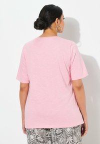 Camiseta de manga corta color rosa claro con un ajuste relajado y cuello redondo, hecha de una tela suave y texturizada. Llevada con pantalones grises con estampado.