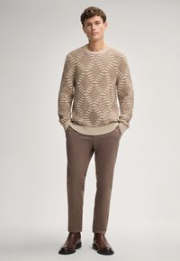 Beiger Strickpullover mit geometrischem Muster, kombiniert mit braunen Slim-Fit-Hosen und dunkelbraunen Stiefeletten. Einfarbiges Hintergrund.