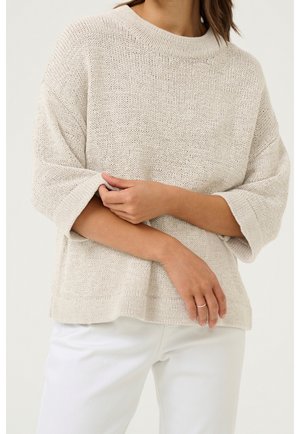 Femme portant un pull en maille beige ample avec des manches jusqu’au coude et un pantalon blanc, debout, les bras croisés à la taille.