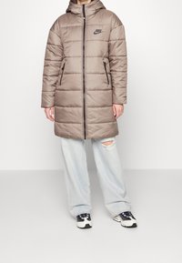 Long, puffy, brown jacket with a hood, featuring horizontal stitching, a front zipper, and black dual side pockets. Logo on chest.