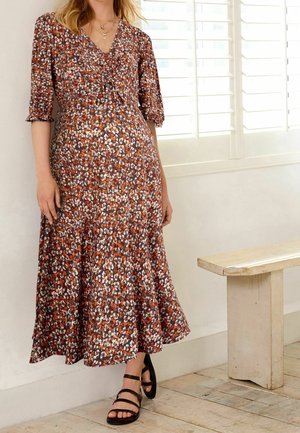 Žena v květinových midi šatech rezavé barvy s 3/4 rukávy a černých páskových sandálech stojící u bílých okenních žaluzií.