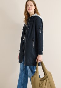 Jeune femme portant un manteau à capuche noir et un jean bleu, tenant un grand sac fourre-tout vert olive sur un fond beige uni.