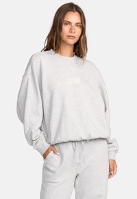 Světlé šedé oversized mikina vyrobená z měkkého materiálu, s kulatým výstřihem, dlouhými rukávy a subtilním logem značky na hrudi.