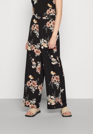Zwarte wijde broek met een bloemenpatroon in tinten roze, crème en groen. Gecombineerd met beige platte sandalen.