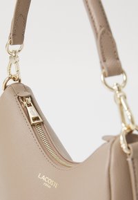Beige textured leather Lacoste handbag with gold zipper, gold zipper pull featuring crocodile logo, and gold hardware on adjustable strap.