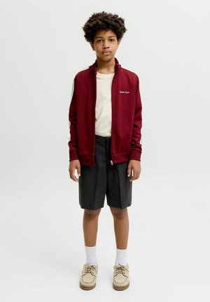 Garçon debout portant une veste zippée bordeaux, une chemise crème, un short noir, des chaussettes blanches et des chaussures beige sur un fond clair uni.