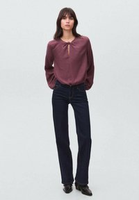 Burgunderbluse mit einem Schlüssel-Ausschnitt und langen Ärmeln, kombiniert mit dunkeltürkisen Weitbeinjeans und schwarzen Stiefeletten.