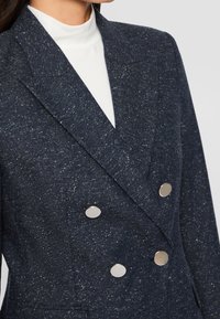 Veste croisée bleu foncé tachetée avec de gros boutons argentés portée sur un haut blanc à col montant.