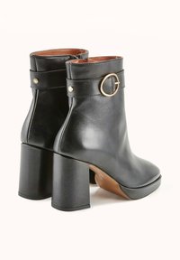 Bottines en cuir noir avec un talon épais, dotées d'un accent circulaire doré sur la sangle et d'une texture lisse. Design élégant et moderne.