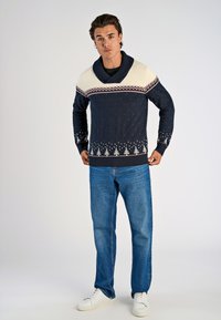 Granatowy sweter dzianinowy z kremowym kołnierzem w stylu szalowym, geometrycznymi wzorami w kolorach czerwonym i białym oraz akcentami w postaci śnieżynek na dole. W komplecie z niebieskimi dżinsami.